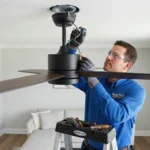 Electrician on a step ladder wiring a black ceiling fan into a ceiling electrical box in a living room