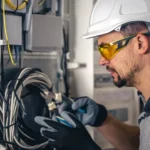 Electrician in a hard hat and safety glasses inspecting wiring inside a residential electrical panel