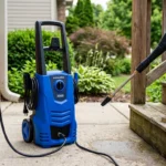 Electric pressure washer plugged into an outdoor outlet on a brick wall while a gloved hand holds the spray wand