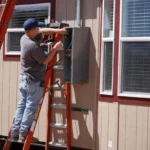 Electrician on a ladder opening an exterior electrical panel on the side of a house