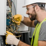 Electrician in a hard hat, safety glasses and hi-vis vest working inside an open electrical distribution panel