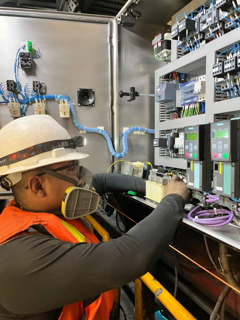 Technician working inside an industrial electrical control cabinet containing variable-frequency drives and terminal blocks